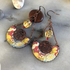 May include: A pair of long, dangling earrings with a crescent moon shape. The earrings are made of metal and have a floral pattern with red, yellow, and white colors. The earrings have a copper-colored chain with small, round beads.