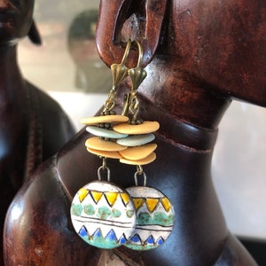 Pu&ograve; includere: Un paio di orecchini in ceramica con un motivo geometrico in tonalit&agrave; di blu, giallo e verde. Gli orecchini sono sospesi da ganci dorati e presentano una pila di perline in ceramica beige e blu chiaro.