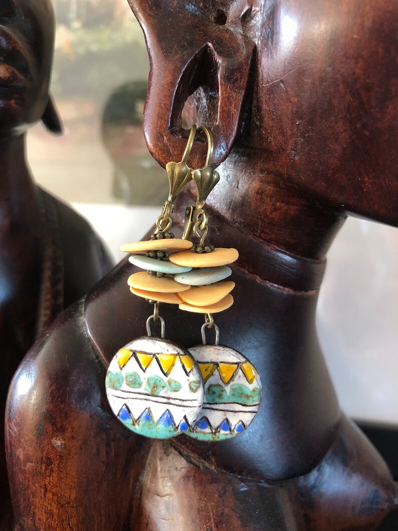 Pu&ograve; includere: Un paio di orecchini in ceramica con un motivo geometrico in tonalit&agrave; di blu, giallo e verde. Gli orecchini sono sospesi da ganci dorati e presentano una pila di perline in ceramica beige e blu chiaro.