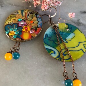 K&ouml;nnte beinhalten: Ein Paar handgefertigte Ohrringe. Einer ist rund mit einem floralen Design und bunten Edelsteinen. Der andere ist oval mit einem blau-gelben Muster. Beide haben Kupferketten und h&auml;ngende Perlen in T&uuml;rkis und Gelb.