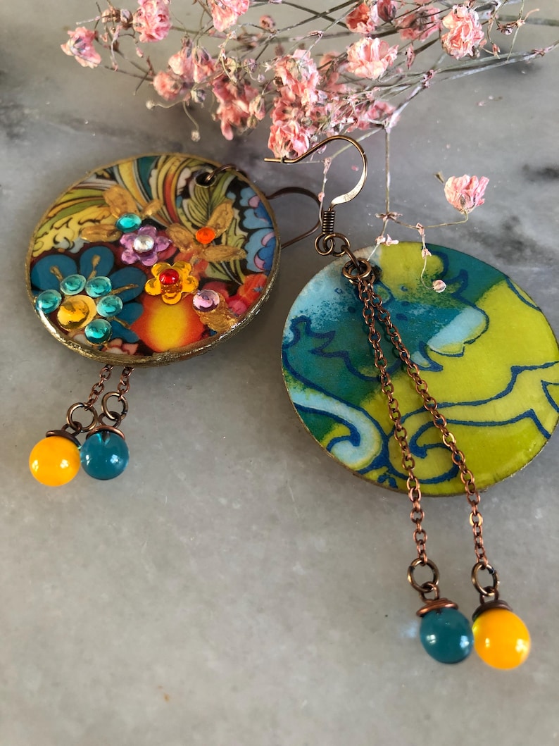 K&ouml;nnte beinhalten: Ein Paar handgefertigte Ohrringe. Einer ist rund mit einem floralen Design und bunten Edelsteinen. Der andere ist oval mit einem blau-gelben Muster. Beide haben Kupferketten und h&auml;ngende Perlen in T&uuml;rkis und Gelb.
