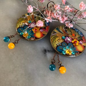 K&ouml;nnte beinhalten: Ein Paar runde, farbenfrohe Ohrringe mit aufwendigen Blumenmustern und h&auml;ngenden Perlen. Die Ohrringe weisen eine Mischung aus Blau-, Gelb- und Oranget&ouml;nen auf, akzentuiert mit kleinen Edelsteinen. Die h&auml;ngenden Perlen sind t&uuml;rkis und gelb. Zarte rosa Bl&uuml;ten sind im Hintergrund.