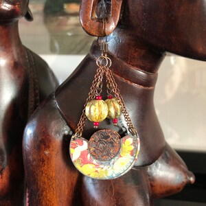 May include: A pair of earrings with a crescent moon shaped pendant featuring a floral design in shades of red, yellow, and white. The pendant is attached to a chain with two small gold beads and red beads.