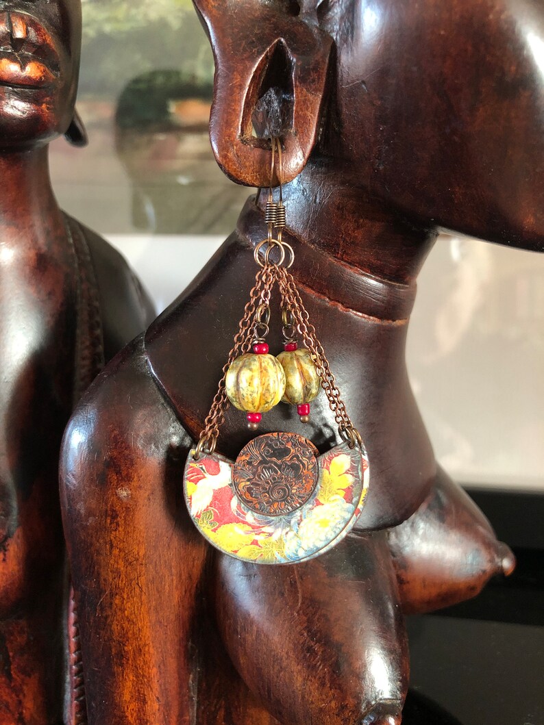 May include: A pair of earrings with a crescent moon shaped pendant featuring a floral design in shades of red, yellow, and white. The pendant is attached to a chain with two small gold beads and red beads.