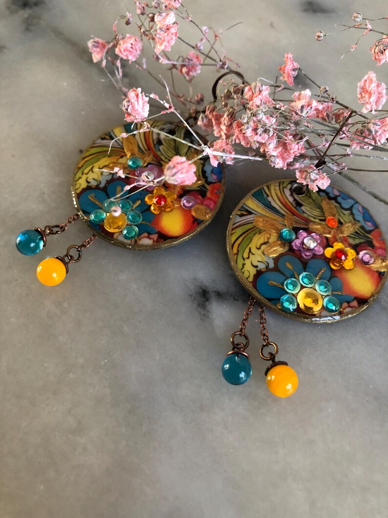 K&ouml;nnte beinhalten: Ein Paar runde, farbenfrohe Ohrringe mit aufwendigen Blumenmustern und h&auml;ngenden Perlen. Die Ohrringe weisen eine Mischung aus Blau-, Gelb- und Oranget&ouml;nen auf, akzentuiert mit kleinen Edelsteinen. Die h&auml;ngenden Perlen sind t&uuml;rkis und gelb. Zarte rosa Bl&uuml;ten sind im Hintergrund.