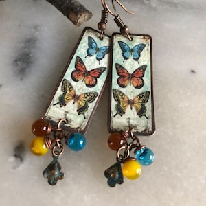 Puede incluir: Un par de pendientes colgantes con un dije rectangular de cobre con un diseño de mariposa vintage. Los dijes están acentuados con cuentas de vidrio azul, amarillo y naranja.