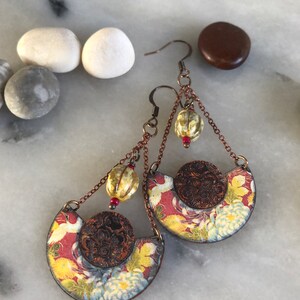 May include: A pair of dangle earrings with a floral pattern and a copper chain. The earrings feature a round, copper-colored charm with a floral design and a small, white bead.