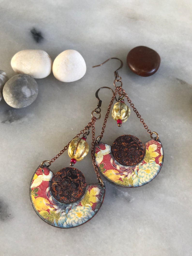 May include: A pair of dangle earrings with a floral pattern and a copper chain. The earrings feature a round, copper-colored charm with a floral design and a small, white bead.