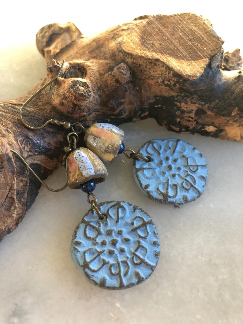 Op de afbeelding: Een paar blauwe keramische oorbellen met een getextureerd ontwerp. De oorbellen hangen aan een bruine tak. Elke oorbel heeft een kleine, ronde, veelkleurige keramische kraal.
