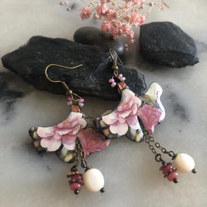 Op de afbeelding: Een paar hangende oorbellen met waaiervormige onderdelen met een roze en wit bloemenpatroon. Elke oorbel heeft een bronskleurige haak, kleine roze kralen en twee bungelende kettinkjes die eindigen in een ronde crèmekleurige en een kleine roze kraal.