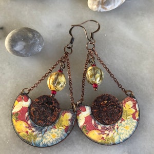 May include: A pair of dangle earrings with a copper finish. The earrings feature a half-circle design with a floral pattern and a small copper disc in the center. The earrings are accented with three small, round, cream-colored beads and red beads.