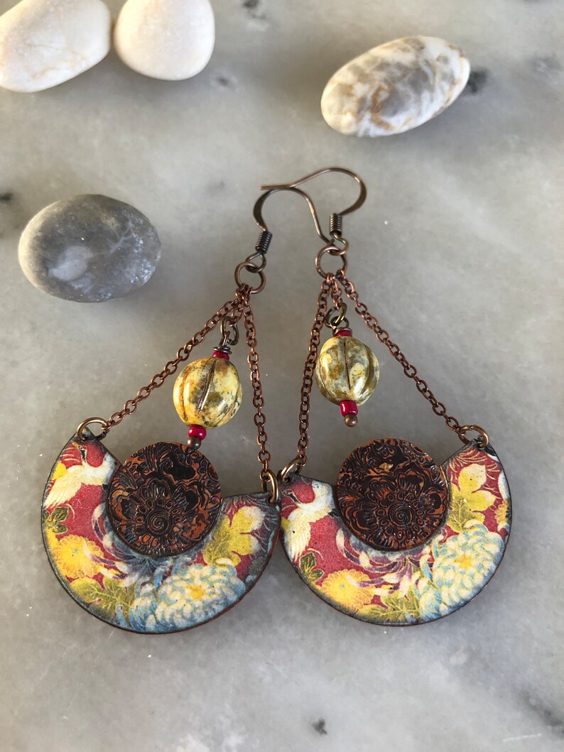 May include: A pair of dangle earrings with a copper finish. The earrings feature a half-circle design with a floral pattern and a small copper disc in the center. The earrings are accented with three small, round, cream-colored beads and red beads.