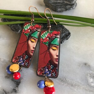 May include: Copper-colored dangle earrings featuring a rectangular pendant with a woman's portrait in a traditional headdress. The earrings have red, blue, and gold beads. The background is red.