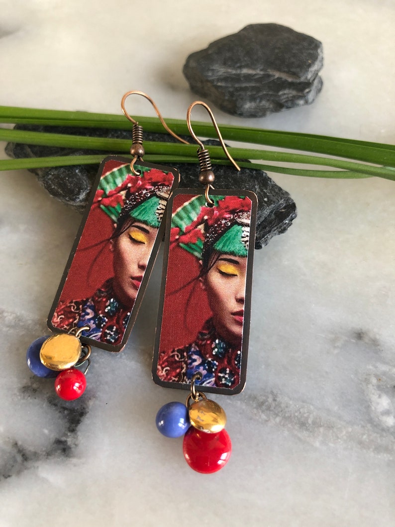 May include: Copper-colored dangle earrings featuring a rectangular pendant with a woman's portrait in a traditional headdress. The earrings have red, blue, and gold beads. The background is red.