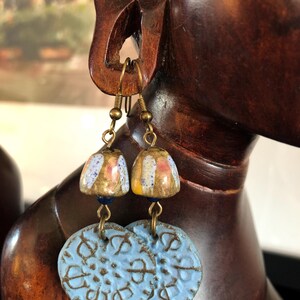 Op de afbeelding: Een paar oorhangers met blauwe en witte keramische schijven en kleine, kleurrijke, keramische kralen. De oorhangers hebben goudkleurige metalen haken.