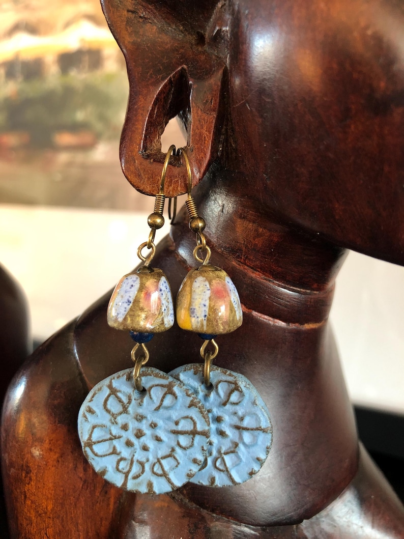 Op de afbeelding: Een paar oorhangers met blauwe en witte keramische schijven en kleine, kleurrijke, keramische kralen. De oorhangers hebben goudkleurige metalen haken.