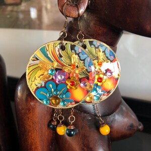 K&ouml;nnte beinhalten: Bunte, runde Ohrringe mit floralem Design und goldenen Akzenten. Die Ohrringe zeigen blaue, lila und rote Edelsteine mit h&auml;ngenden gelben und schwarzen Perlen. Die Ohrringe h&auml;ngen an einer Holzskulptur.