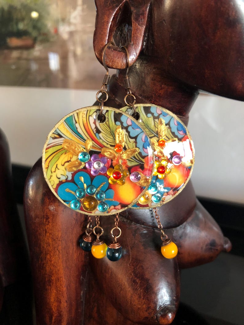 K&ouml;nnte beinhalten: Bunte, runde Ohrringe mit floralem Design und goldenen Akzenten. Die Ohrringe zeigen blaue, lila und rote Edelsteine mit h&auml;ngenden gelben und schwarzen Perlen. Die Ohrringe h&auml;ngen an einer Holzskulptur.