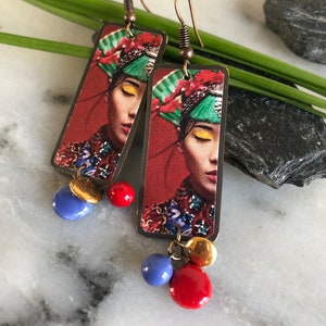May include: Copper-toned dangle earrings. Each earring features a rectangular pendant with a woman's portrait, red background, and colorful accents. Dangling from each pendant are blue, red, and gold-toned beads. The earrings have a hook closure.