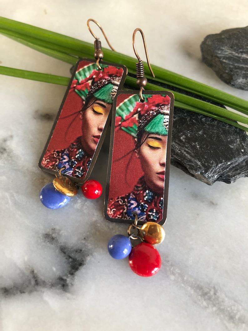 May include: Copper-toned dangle earrings. Each earring features a rectangular pendant with a woman's portrait, red background, and colorful accents. Dangling from each pendant are blue, red, and gold-toned beads. The earrings have a hook closure.