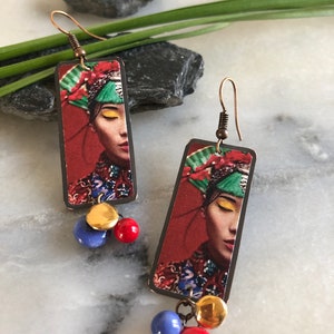 May include: A pair of rectangular earrings featuring a woman's face with a red background. The earrings have copper-colored hooks and are adorned with gold, blue, and red beads. The woman in the image is wearing a colorful headdress.