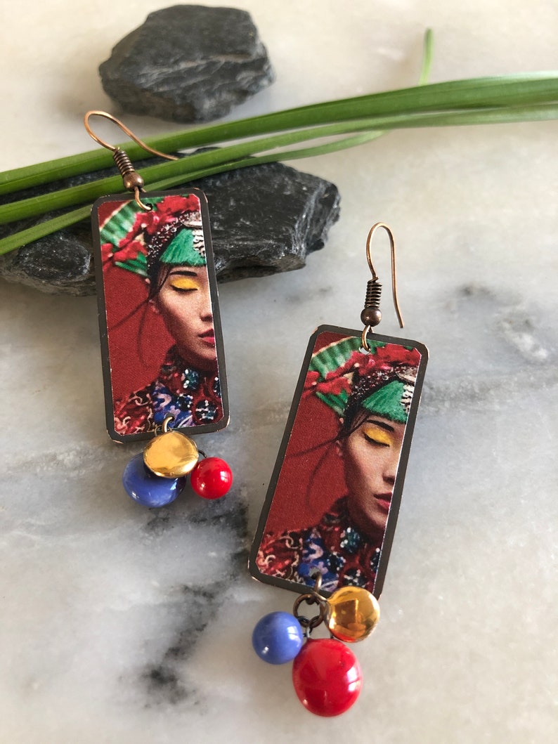 May include: A pair of rectangular earrings featuring a woman's face with a red background. The earrings have copper-colored hooks and are adorned with gold, blue, and red beads. The woman in the image is wearing a colorful headdress.
