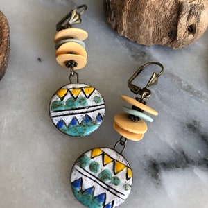 Pu&ograve; includere: Un paio di orecchini pendenti con un ciondolo rotondo in ceramica con un motivo geometrico in tonalit&agrave; di blu, giallo e bianco. I ciondoli sono sospesi da una pila di perline beige e blu chiaro.