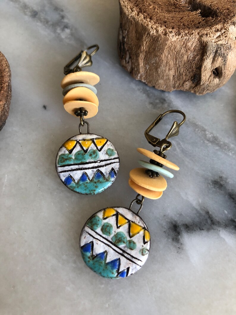 Pu&ograve; includere: Un paio di orecchini pendenti con un ciondolo rotondo in ceramica con un motivo geometrico in tonalit&agrave; di blu, giallo e bianco. I ciondoli sono sospesi da una pila di perline beige e blu chiaro.