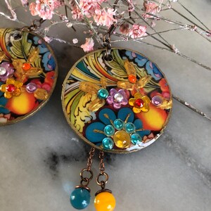K&ouml;nnte beinhalten: Bunte, runde Ohrringe mit floralen Designs und Schmuckakzenten. Die Ohrringe weisen eine Mischung aus Blau, Gelb, Orange und Lila auf. Jeder Ohrring hat einen goldfarbenen Rand und eine baumelnde blaue und gelbe Perle.