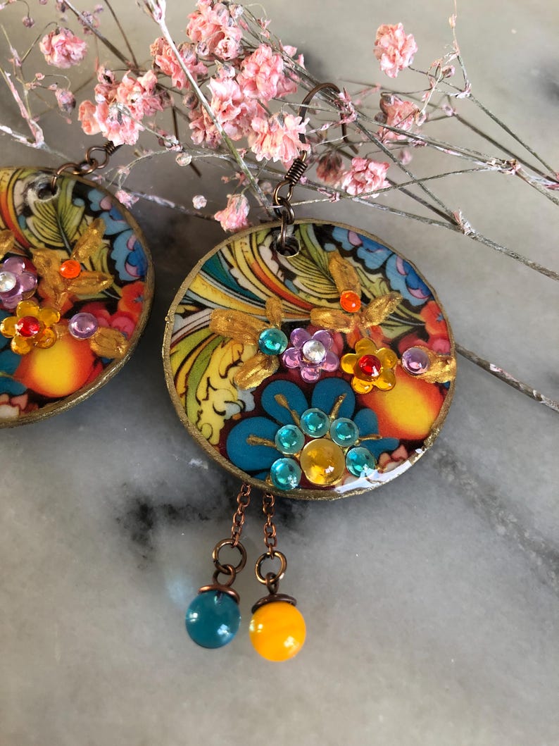 K&ouml;nnte beinhalten: Bunte, runde Ohrringe mit floralen Designs und Schmuckakzenten. Die Ohrringe weisen eine Mischung aus Blau, Gelb, Orange und Lila auf. Jeder Ohrring hat einen goldfarbenen Rand und eine baumelnde blaue und gelbe Perle.