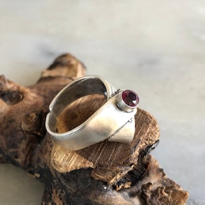 May include: A silver ring with a dark red gemstone set in the center. The ring is on a piece of wood.