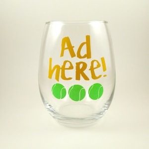 May include: A clear glass wine glass with the words "Ad here!" in gold lettering and three green tennis balls printed on the front.