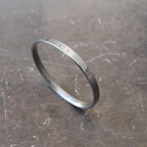 Könnte beinhalten: Ein dünner, polierter silberfarbener Metallring mit einer glatten, abgerundeten Außenfläche und einem einfachen, kreisförmigen Banddesign. Der Ring verfügt über eine reflektierende Metalloberfläche, die das Licht einfängt und ein klares, einheitliches Erscheinungsbild zeigt.