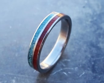 May include: A metallic band ring featuring two distinct inlaid channels. One channel contains a textured, granular turquoise-colored material, while the adjacent channel consists of a smooth, solid deep red material set within the silver-toned metal.
