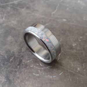May include: A silver-toned metal ring featuring a wide, flat band with a central inlay of crushed white opal material. The inlay displays iridescent flecks of pink and blue light, contrasting against the polished, smooth metallic surface of the ring.