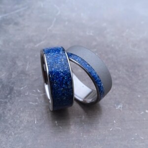 May include: Two rings with a matte grey metal finish. One ring features a wide band of textured, deep blue crushed stone inlay, while the other ring has a thinner, centred inlay of the same blue material circling the band.