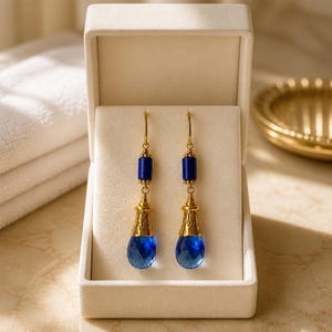 May include: A pair of gold-tone drop earrings featuring a cylindrical dark blue stone bead, a textured gold-tone metal cap, and a faceted teardrop-shaped blue crystal pendant, all presented inside a cream-colored velvet-lined jewelry box.