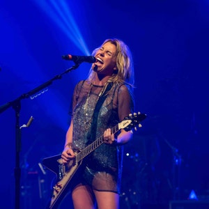 Może przedstawiać: Kobieta w błyszczącej, przezroczystej bluzeczce śpiewa do mikrofonu, grając na czarnej gitarze elektrycznej z białym pickguardem. Na gitarze widnieje napis "Gibson".