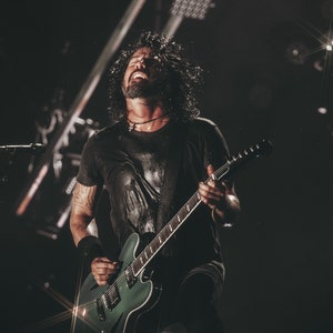 Puede incluir: Un hombre con el pelo largo y oscuro canta en un micrófono mientras toca una guitarra eléctrica verde con una placa de protección negra. Lleva una camiseta negra y pantalones negros. El fondo es oscuro con focos.