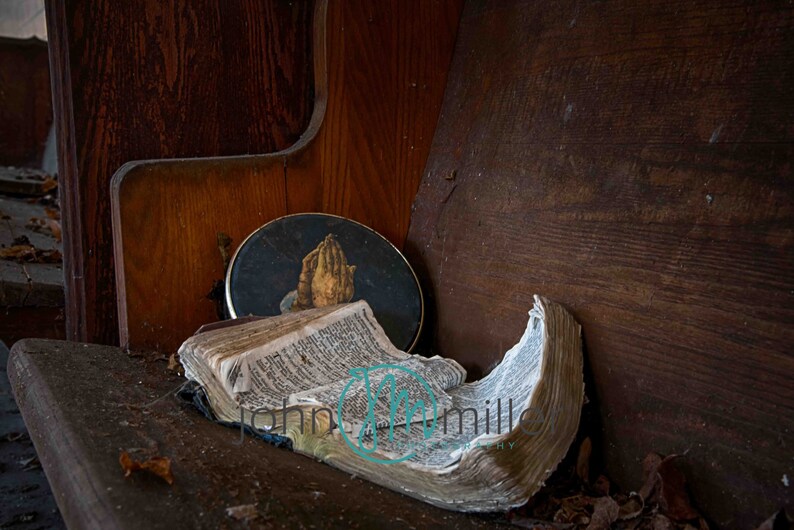 May include: An open, worn bible lies on a wooden pew with a round, dark blue and gold picture of a pair of hands clasped in prayer.