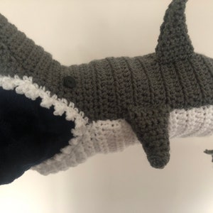May include: Grey and white crocheted shark puppet with a button eye. The shark's mouth is open and has white teeth.