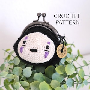 May include: Crochet pattern for a No Face coin purse. The purse is white with black details and purple accents. The purse is sitting on a bed of green leaves. The text "CROCHET PATTERN" is visible in the image.