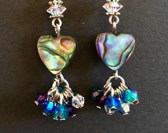 May include: A pair of dangle earrings featuring heart-shaped abalone shell beads with iridescent green and purple tones. Each earring includes a clear faceted crystal bead above the heart and a cluster of small blue, purple, and clear crystal beads.