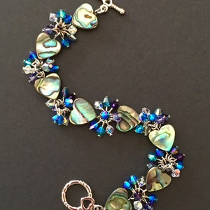 May include: A bracelet featuring heart-shaped abalone shell charms interspersed with clusters of faceted blue, purple, and clear crystal beads. The piece is secured with a silver-toned metal toggle clasp shaped like a heart at one end.