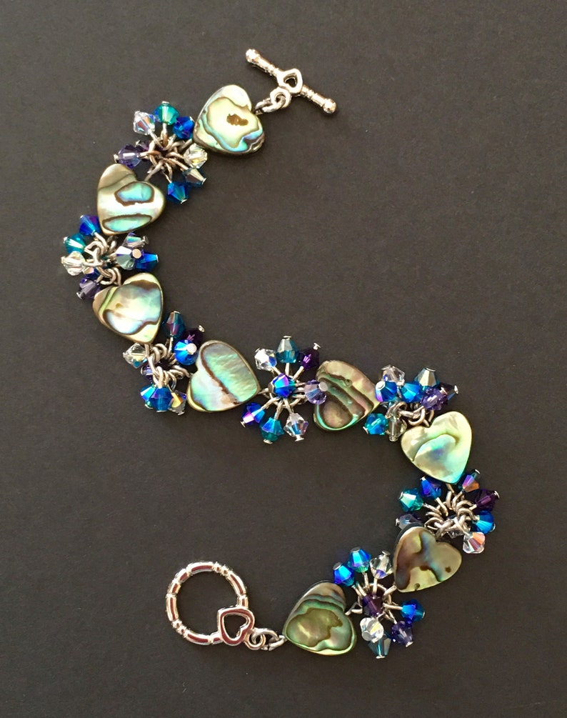 May include: A bracelet featuring heart-shaped abalone shell charms interspersed with clusters of faceted blue, purple, and clear crystal beads. The piece is secured with a silver-toned metal toggle clasp shaped like a heart at one end.