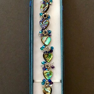 May include: A bracelet featuring a series of heart-shaped abalone shell beads in iridescent green, blue, and brown tones. The hearts are linked by silver-toned metal rings and accented with small, faceted crystal beads in shades of blue and clear.