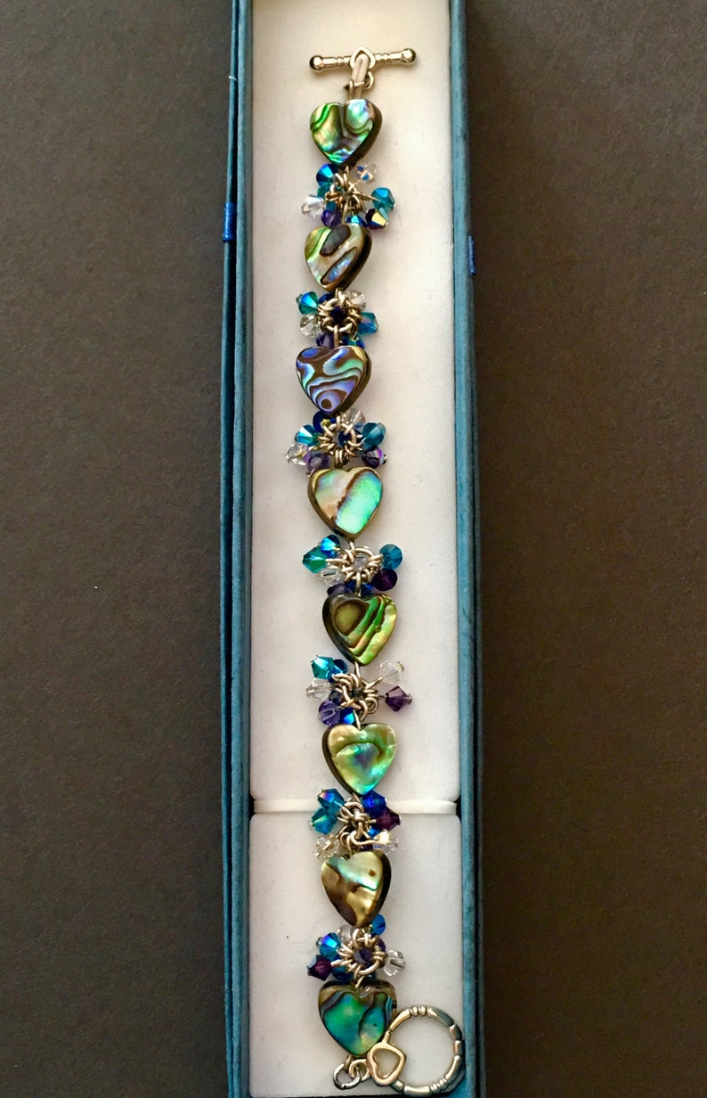 May include: A bracelet featuring a series of heart-shaped abalone shell beads in iridescent green, blue, and brown tones. The hearts are linked by silver-toned metal rings and accented with small, faceted crystal beads in shades of blue and clear.