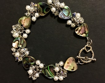 May include: A charm bracelet featuring iridescent abalone shell heart-shaped beads, small white round pearls, and clear faceted crystal beads linked together with silver-toned metal chains and a heart-shaped toggle clasp closure.