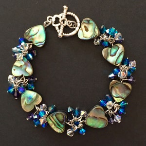 May include: A bracelet featuring heart-shaped abalone shell beads interspersed with clusters of faceted blue and clear crystal beads. The piece is secured with a silver-toned metal toggle clasp shaped like a heart, connecting the ends of the bracelet.