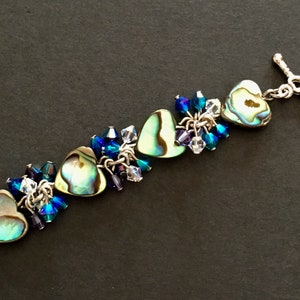 May include: A bracelet featuring three heart-shaped abalone shell charms interspersed with clusters of faceted blue, purple, and clear crystal beads. The jewelry piece is finished with a silver-toned metal toggle clasp shaped like a small heart.